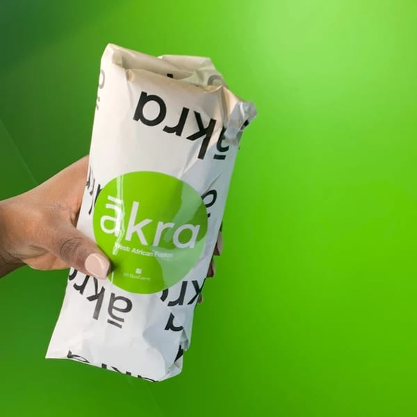A hand holds a white ākra Eatery takeout bag with a green logo against a bright green background.
