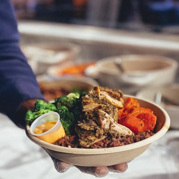 Gloved hands hold an African-inspired bowl with chicken, broccoli, sweet potatoes, rice, and sauce.
